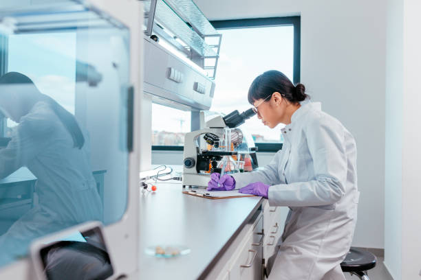 A female scientist is sitting in her lab and looking trough the microscope, she is writing down notes about her discoveries after looking at samples